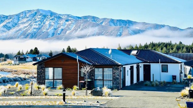 Een modern huis met een stenen gevel en grote ramen, tegen een achtergrond van besneeuwde bergen en een bebost gebied.