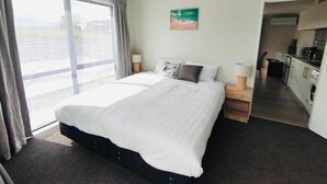 A hotel room with a large bed, a view through the window, a painting on the wall, and a kitchenette in the adjacent room. - Stellar A- Sweet unit in Lake Tekapo (Lake Tekpao)