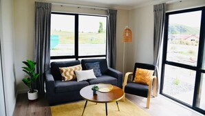 A modern living room with a black sofa, a wooden coffee table, and a view of a green landscape through large windows. - Stellar A- Sweet unit in Lake Tekapo (Lake Tekpao)