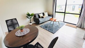 Ein modernes Wohnzimmer mit einem runden Esstisch, zwei schwarzen Stühlen, einem grauen Sofa mit Kissen und einem kleinen orangefarbenen Beistelltisch. Ein großes Fenster mit Vorhängen rundet das Bild ab.