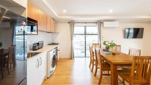 Cocina moderna con horno empotrado, microondas y lavadora. Cuenta con una zona de comedor con mesa y sillas de madera, y un amplio ventanal con vistas al exterior.