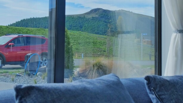 A room with a large window overlooking a red car and a green landscape with hills. - Starry Heaven 2bdr2ba (Lake Tekapo)