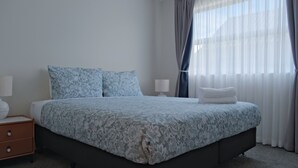 A neatly made bed with a floral-patterned bedspread and pillows, a bedside table with a lamp, and a window with sheer curtains. - Starry Heaven 3bdr3ba (Lake Tekapo)