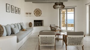 A modern living room with a sofa, two wicker chairs, a fireplace, and a view of the sea. - Luxury 5BDR Retreat in Mykonos (Mikonos)