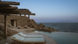 A stone building with a wooden pergola, a pool, and lounge chairs overlooking the sea. - Luxury 5BDR Retreat in Mykonos (Mikonos)