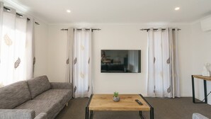 Een moderne woonkamer met een grijze bank, een houten salontafel met een plant, en een aan de muur bevestigde televisie.