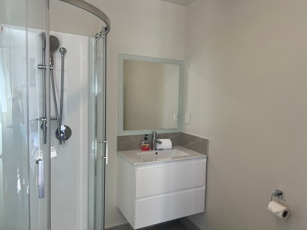 Un baño moderno con cabina de ducha de vidrio, tocador blanco con espejo y dispensador de papel higiénico en la pared.