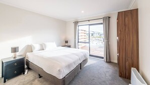 A bedroom with a large bed, a nightstand with a lamp, a wooden wardrobe, and a sliding glass door to a balcony. - Mirror Lake 2 Bedroom A (Lake Tekapo)