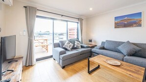A modern living room with a grey sofa, a wooden coffee table, and a large window offering a view of the city. - Mirror Lake 2 Bedroom A (Lake Tekapo)