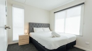 Ein modernes Schlafzimmer mit grauem, gepolstertem Kopfteil, einem Holznachttisch, einer weißen Bettdecke und einem großen Fenster mit Jalousien.