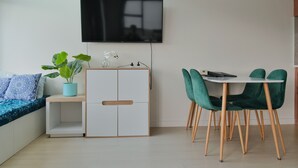 Un espace de vie moderne avec un téléviseur fixé au mur, un canapé agrémenté de coussins à motifs, une petite table avec une plante et un coin repas avec des chaises vertes.