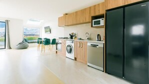 Une cuisine moderne avec des meubles en bois, un réfrigérateur noir, un four à micro-ondes, un lave-linge et un lave-vaisselle. On y trouve deux chaises vertes et un fauteuil en forme de poire de couleur grise, près d’une fenêtre donnant sur l’extérieur.