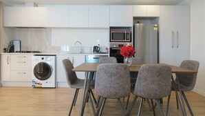 A modern kitchen with white cabinets, a stainless steel refrigerator, a microwave, and a washing machine. There is a dining table with four chairs and a vase with red flowers. - Jeros 2bdr Apartment (Lake Tekapo)