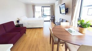 Une chambre d’hôtel comprenant un lit, une table à manger avec des chaises, un canapé, un téléviseur fixé au mur et une fenêtre avec des rideaux.