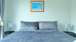 Habitación de hotel con cama, dos mesitas de noche, lámpara, un cuadro en la pared y una ventana con cortinas.