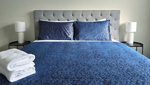 A neatly made bed with a blue patterned bedspread and pillows, two bedside lamps, and a stack of white towels on the left side.