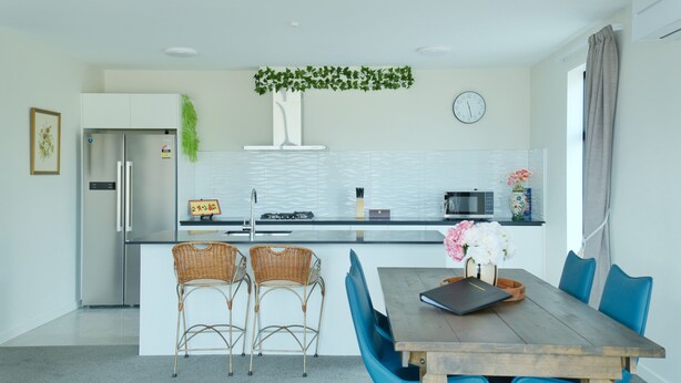 A modern kitchen with a central island, two wicker chairs, a dining table with chairs, a clock, and a window with curtains. - Stargazing Tekapo Apartment C (Lake Tekapo)
