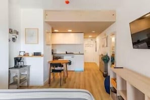 A compact hotel room with a bed, a small desk, a chair, a kitchenette, and a TV.