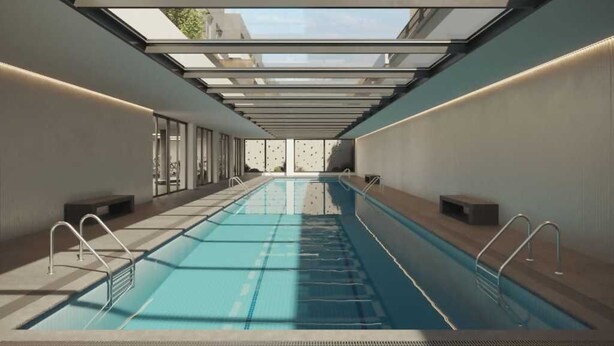 A modern indoor swimming pool with a skylight ceiling, clear poolside seating, and a view of the surrounding buildings.