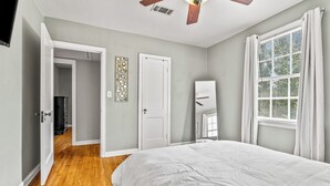 Une chambre à coucher comprenant un lit, un ventilateur de plafond, une fenêtre avec des rideaux et une porte donnant sur une autre pièce.