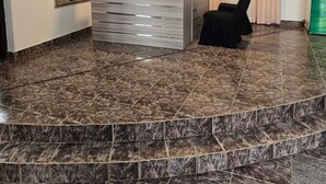 Una zona d'entrada amb sòl de marbre, una taula d'informació, una cadira negra, un mirall gran i una làmpada al sostre.