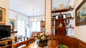 A dining area with a wooden table, chairs, and a vase of flowers. There is a television on a stand to the left.
