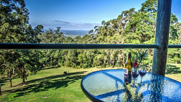 A glass table with a bottle and two glasses on a balcony overlooking a lush landscape.