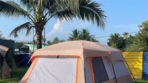 A large, beige tent with a grey floor and orange trim, set up on a grassy area with palm trees and a blue fence in the background. - RAZO VILLAGE INN (Taiping)