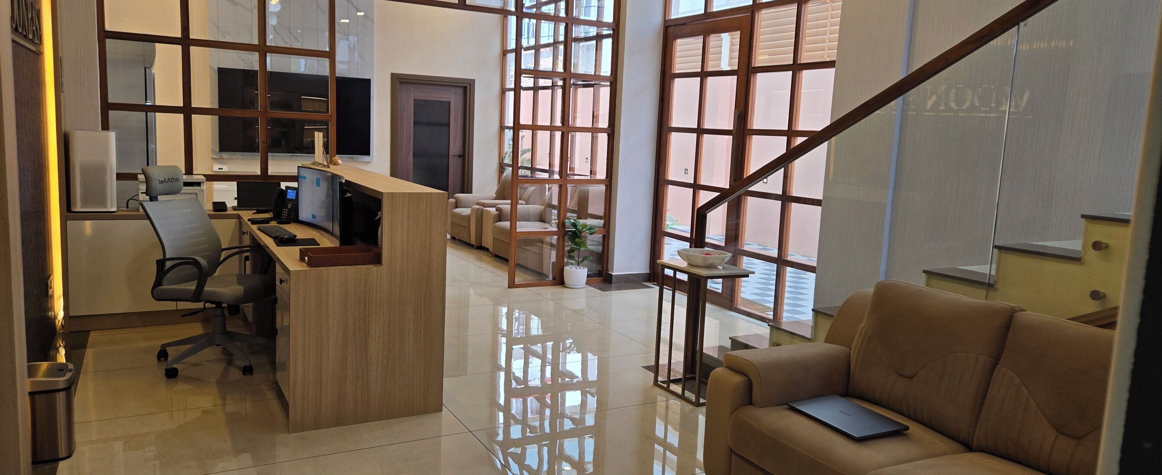 A modern office space with a reception desk, computer, and office chairs. There is a sofa and a plant in the corner.