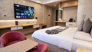 A modern hotel room with a bed, a desk, a chair, a TV mounted on the wall, and a small kitchenette area.
