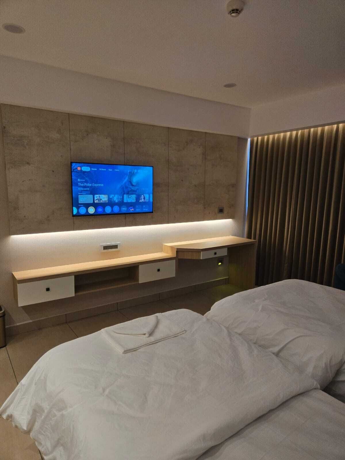 A modern hotel room with a large bed, a flat-screen TV mounted on the wall, and a wooden console table with drawers.