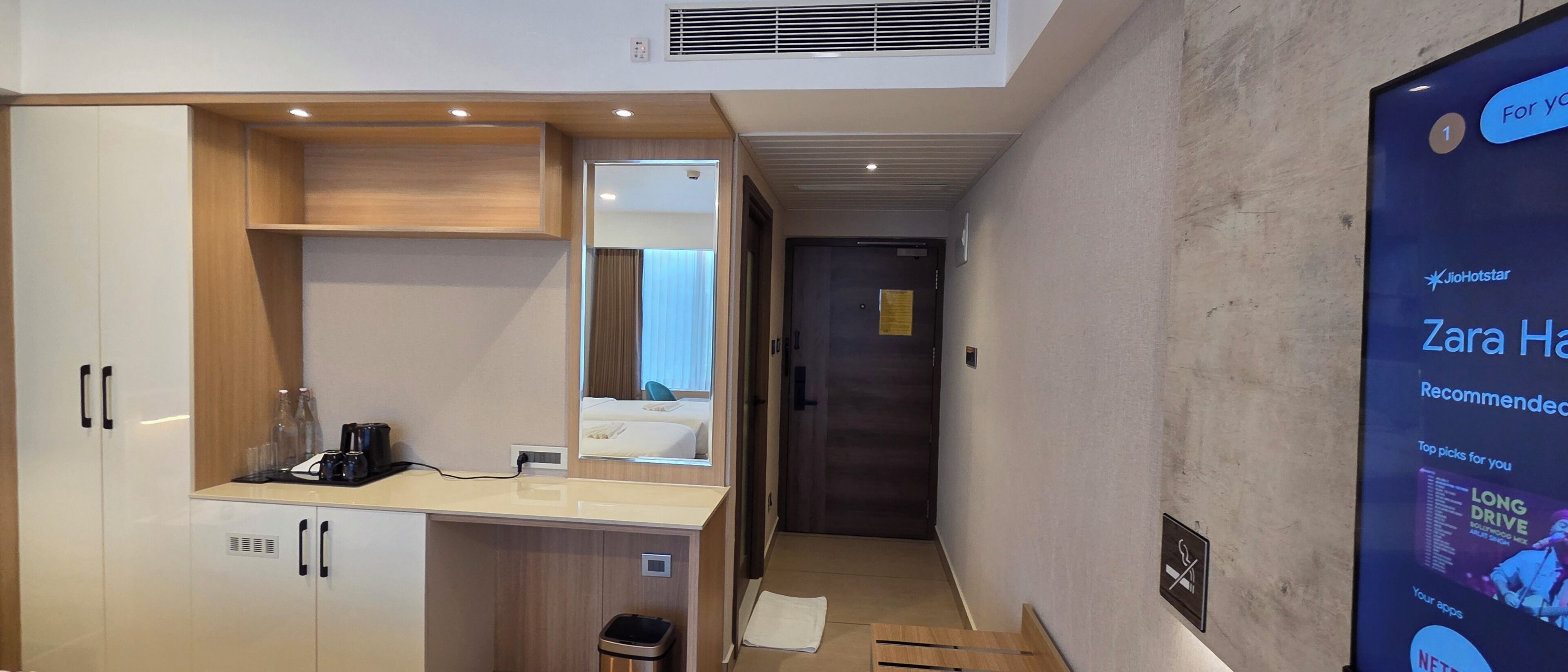 Habitación de hotel con minibar, espejo, mueble de madera, cubo de basura y una pantalla digital mostrando contenido.