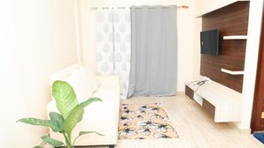 Una habitación pequeña y bien iluminada con un sofá, una planta en maceta, un tapete estampado, un televisor en un estante de madera y una cortina.