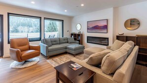 A modern living room with a brown leather armchair, a grey sectional sofa, and a beige sofa. There is a fireplace, a large window with a view of trees, and a round mirror on the wall.