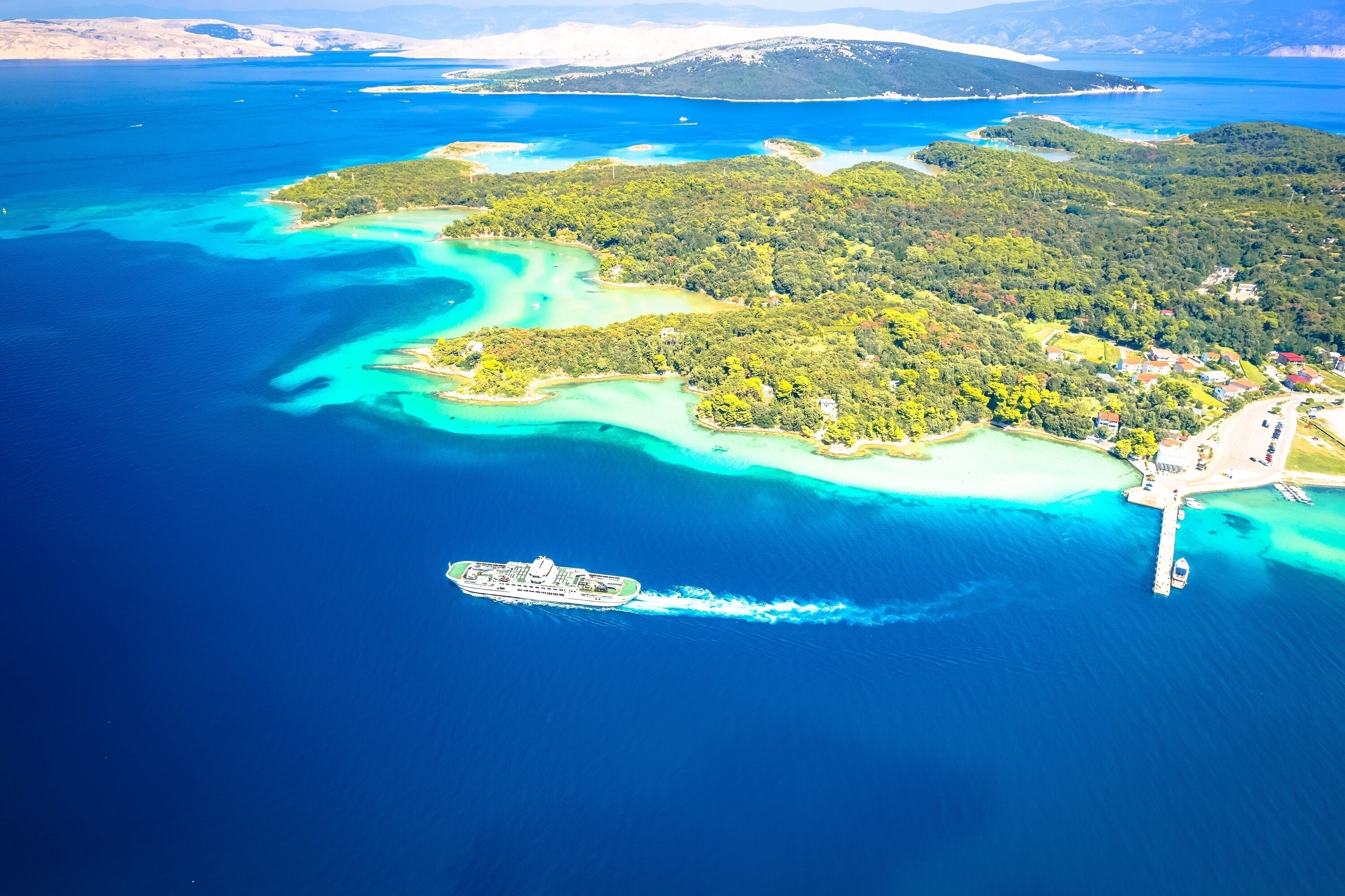 An aerial view of a coastal area with a cruise ship docked near a pier, surrounded by turquoise waters and lush green islands.