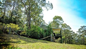 Uma casa com varanda e área coberta, cercada por árvores e um declive gramado.