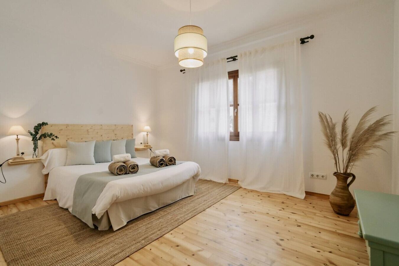 Ett sovrum med en sänggavel i trä, en säng med hoprullade handdukar, sänglampor, ett fönster med tunna gardiner och en vas med pampasgräs.