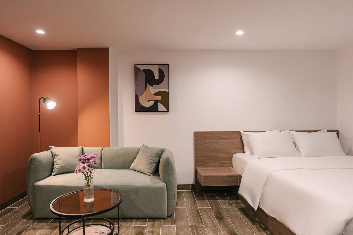 A modern hotel room with a sofa, a round coffee table with a vase of flowers, a bed with white bedding, and a wall-mounted abstract art piece.