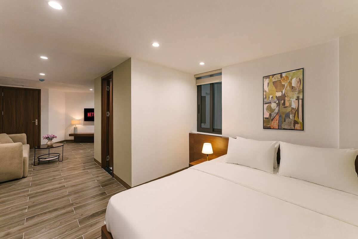 A hotel room with a bed, a bedside table with a lamp, a wooden floor, a sofa, and a painting on the wall.