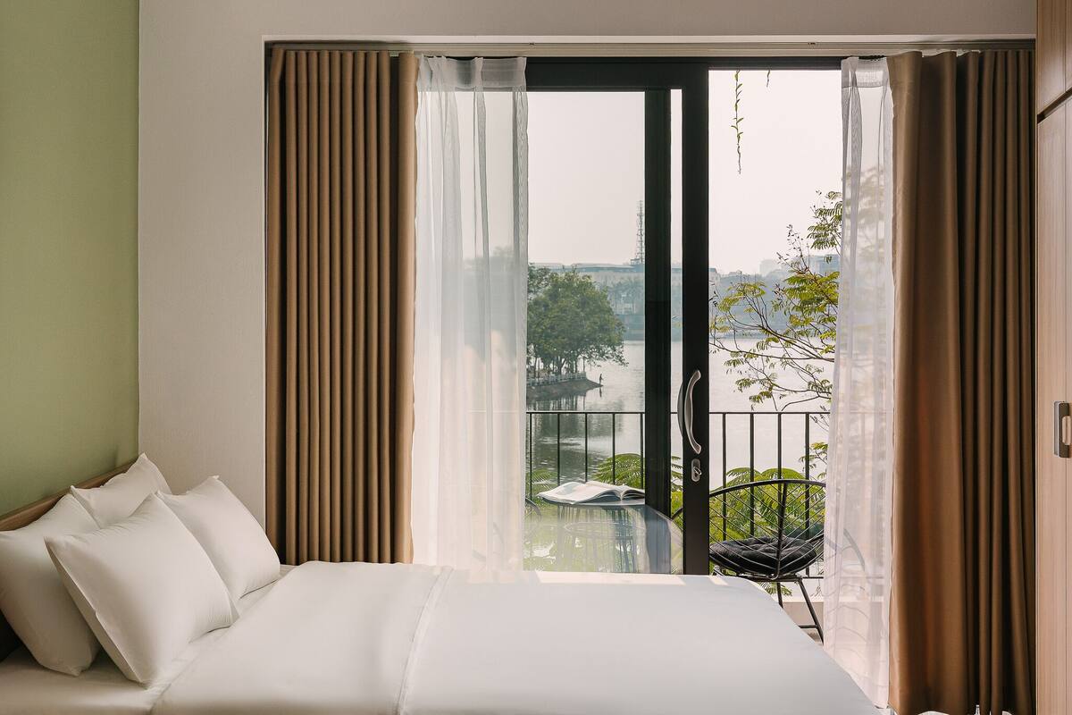 A hotel room with a bed, a view of a balcony with a chair, and a table with a book.