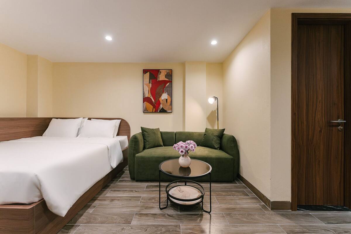 A bedroom with a bed, a sofa, a small table with a vase, and a painting on the wall.