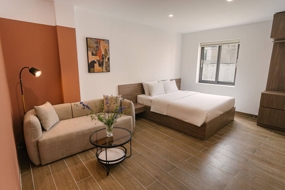 A modern hotel room with a sofa, a bed, a small table with a vase, a floor lamp, and a painting on the wall.