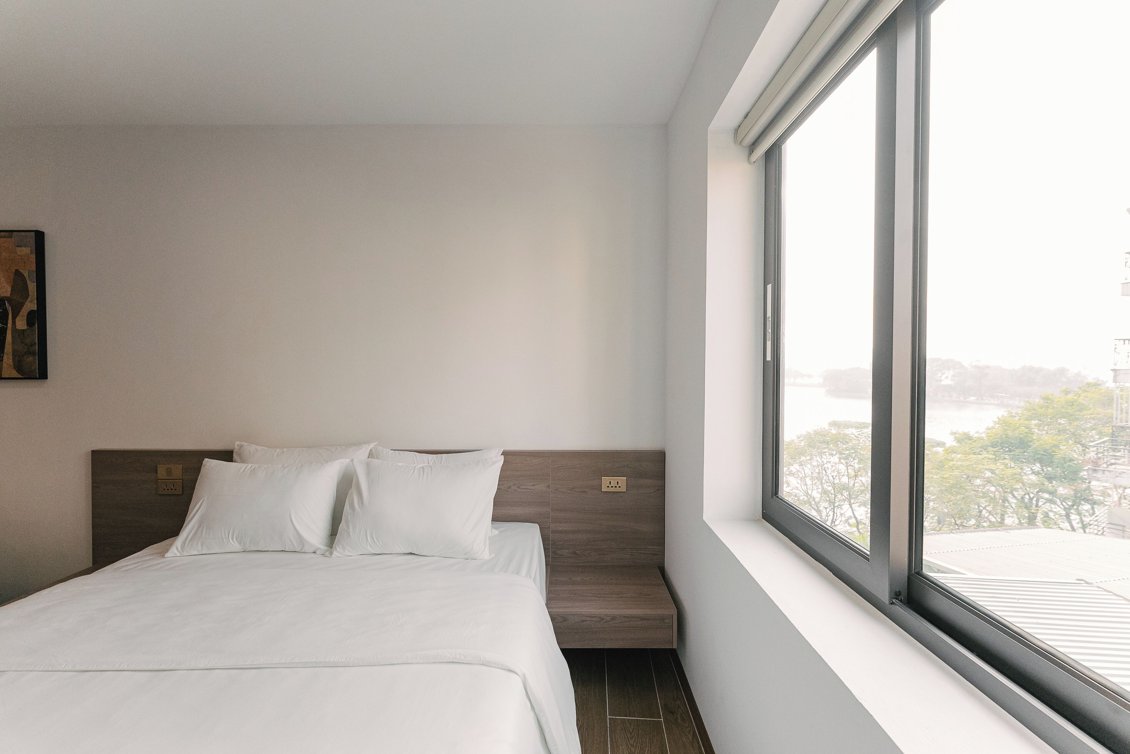 A modern hotel room with a large window, a bed with white linens, and a wooden headboard.