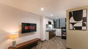 A modern hotel room with a flat-screen TV displaying 'NETFLIX', a wooden bench, a kitchenette, and a large abstract painting on the wall. - An Nguyen Lakeside Residence (Hanoi)