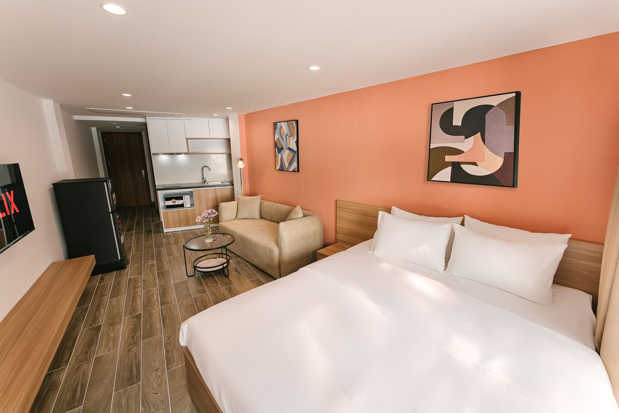 A hotel room with a large bed, a sofa, a small table, and a kitchenette in the background.