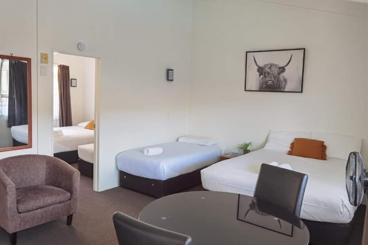 A hotel room with two single beds, a round table with chairs, a sofa, a chair, and a wall-mounted fan.