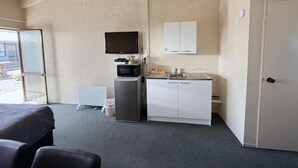 Une chambre d’hôtel avec un lit, une kitchenette équipée d’un four à micro-ondes, d’un évier et de placards. Il y a une porte sur la droite et une fenêtre sur la gauche.