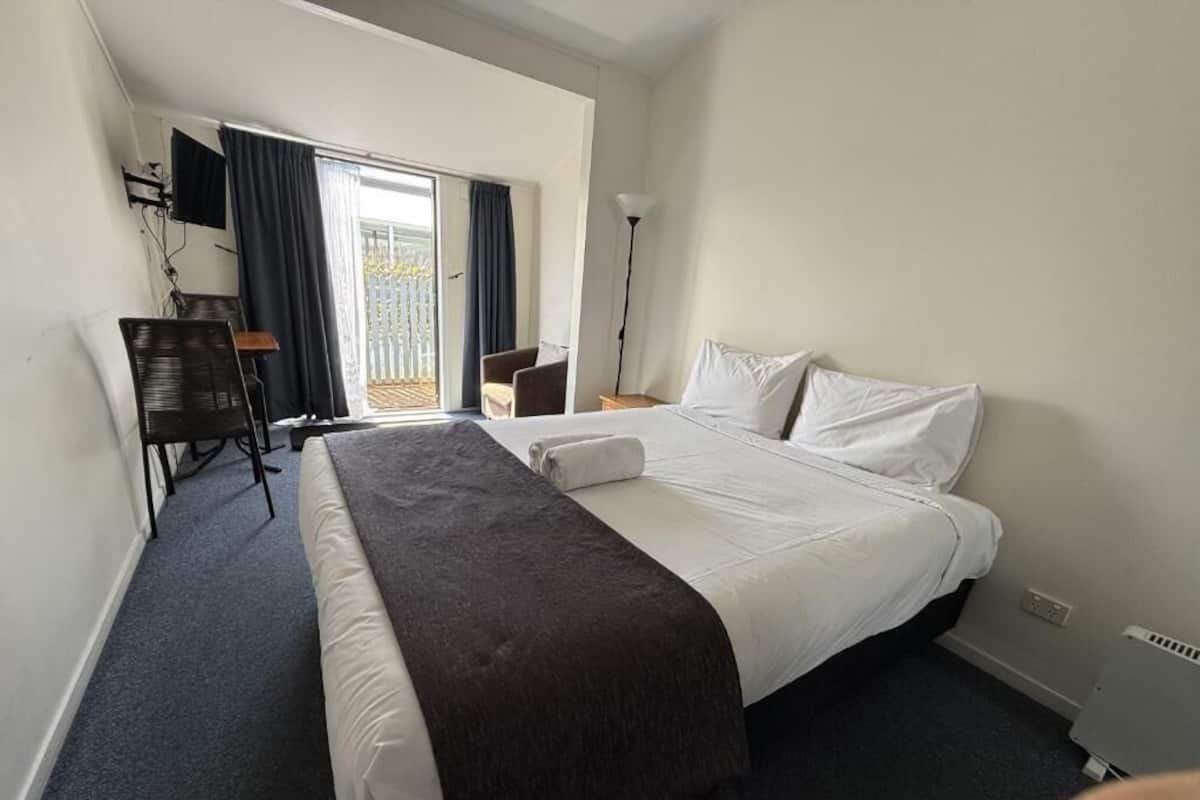 A hotel room with a large bed, a desk with a chair, a TV mounted on the wall, and a window with curtains.