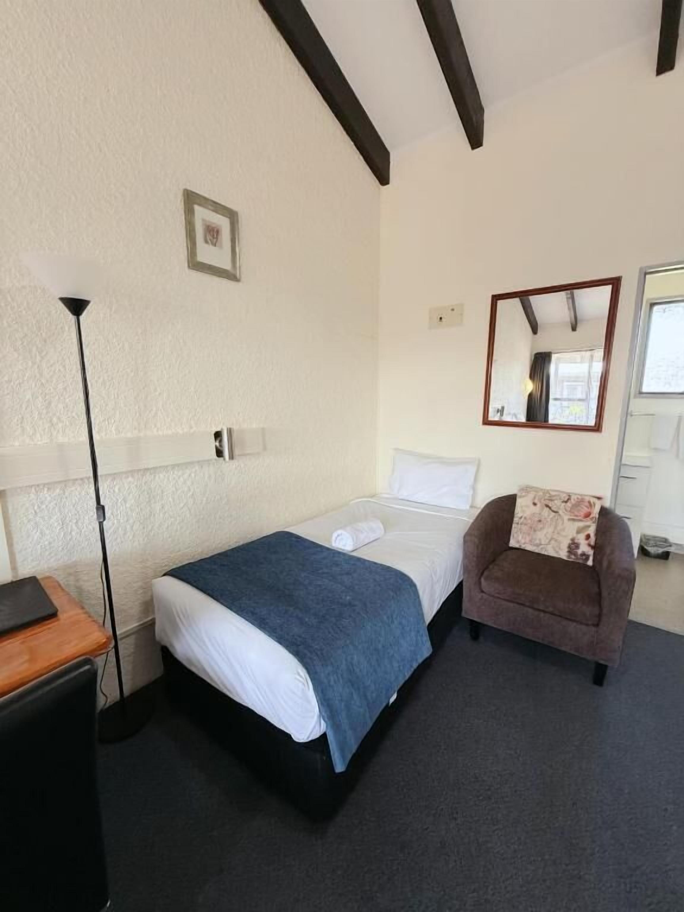 A single bed with a blue blanket, a wooden desk, a brown armchair, a floor lamp, and a mirror on the wall.