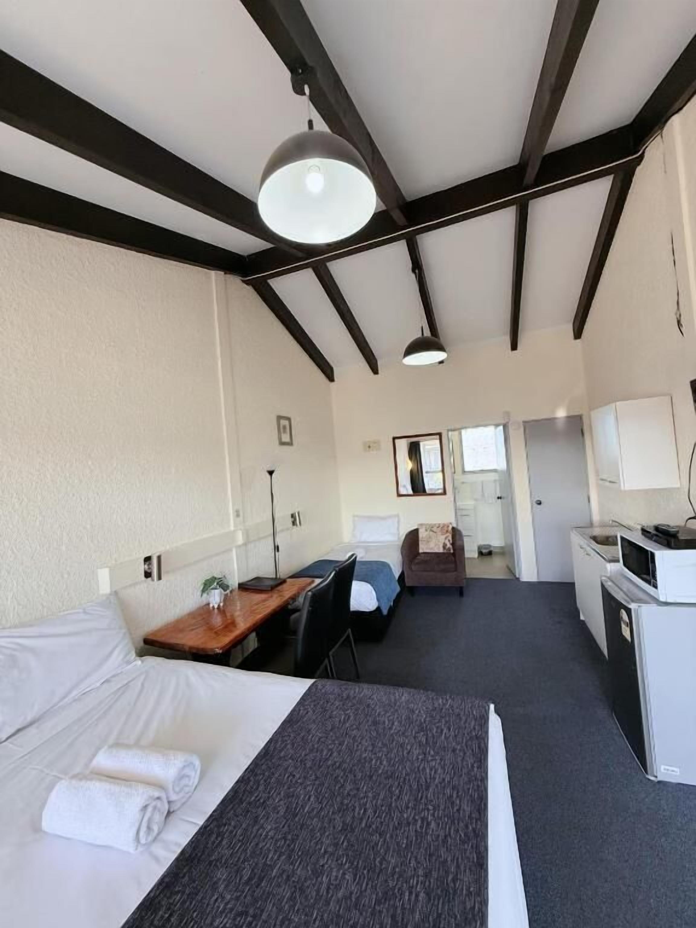 Habitación de hotel con cama, escritorio, silla, kitchenette y baño visible a través de una puerta abierta.